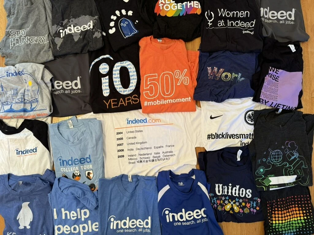 About two dozen Indeed T-shirts laid out on a wood floor, including a tenth-anniversary tee, an indeed.com launch timeline tee, and the College Tour penguins.